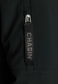 Tela negra con textura suave que presenta un bolsillo con cremallera. Primer plano de una etiqueta con "CHASIN'" grabada en una tira de tela.