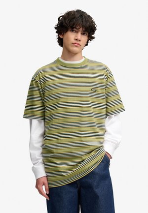 Junger Mann mit kurzen, welligen Haaren, der ein grün-weiß gestreiftes kurzärmeliges Hemd über einem weißen langärmeligen Hemd und blauen Jeans trägt.