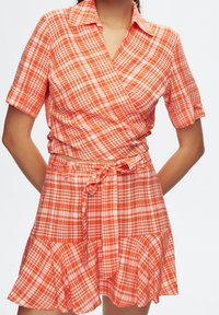 Zavinovací šaty z oranžové kostkované látky s krátkými rukávy, límečkem, zavazováním v pase a rozšířeným lemem. Výrazný kostkovaný vzor v různých velikostech.