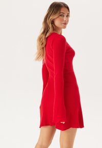 Rode geribbelde jurk met lange mouwen en een uitlopende zoom, met een aansluitende bodice en een gladde textuur. De achterkant heeft een iets verhoogde halslijn.