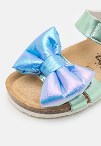 Metallisk grön sandal med justerbar rem, prydd med en stor iriserande blå klänning och en texturerad korksula. Vit gummisula.