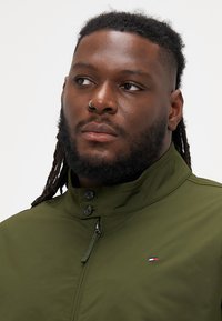 Olivengrøn jakke med høj krave, trykknapper og subtilt logo-detalje. Glat tekstur med et moderne, tætsiddende design.