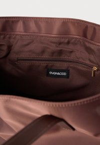 Interior de tecido castanho de uma bolsa com um bolso com fecho e uma etiqueta da marca Even&Odd em preto cosida abaixo do fecho.