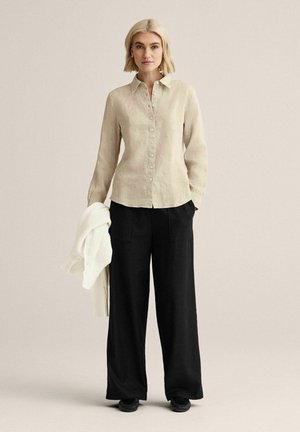 Lyhytukkainen vaaleahiuksinen nainen pukeutunut beigeen nappipaitaan ja mustiin leveisiin housuihin, vasemmassa kädessään valkoinen neuleneuletakki.