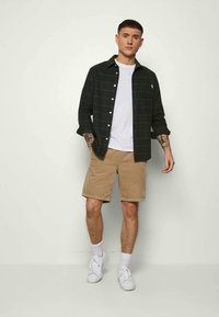 Jovem com cabelo curto, vestindo uma camisa preta de botão sobre uma t-shirt branca, calções bege, meias brancas e ténis brancos, de pé contra uma parede simples.