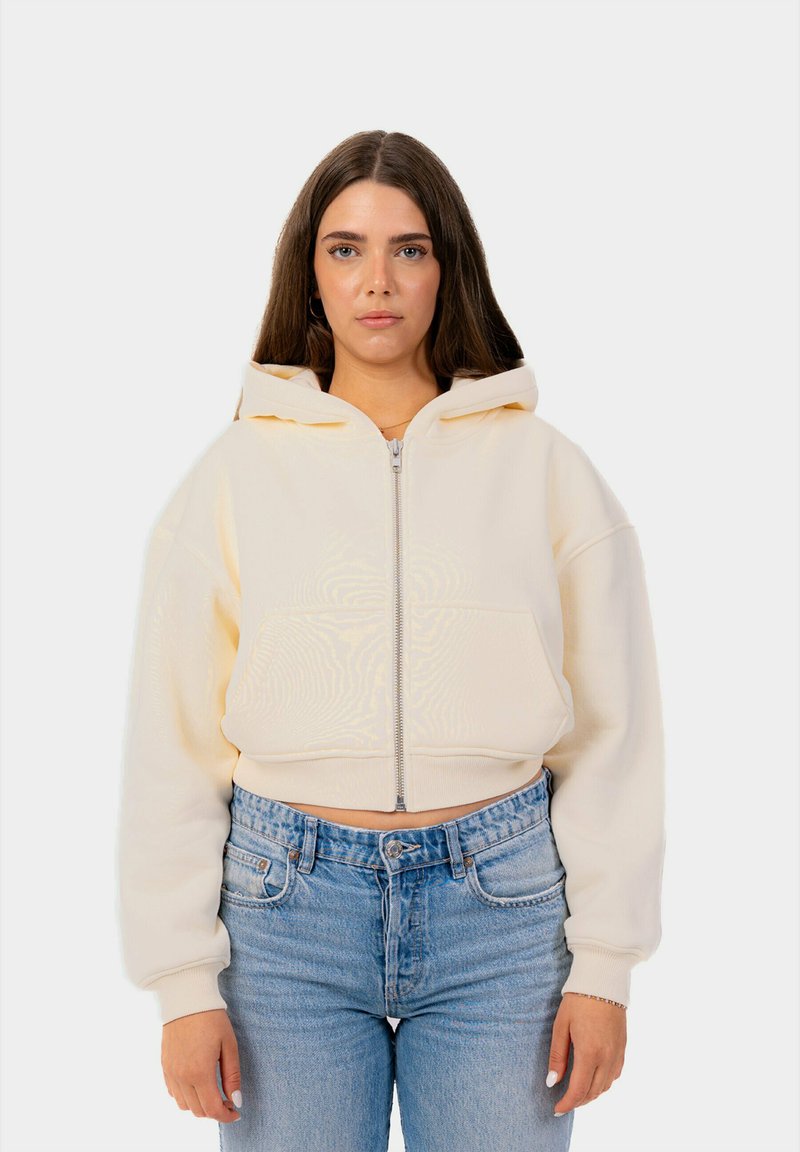 Sudadera recortada en crema con cremallera, capucha y bolsillos frontales, con un fondo liso. Combinada con unos jeans de tiro alto en azul claro.