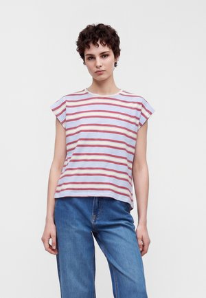 Jeune personne aux cheveux courts et bouclés porte un haut sans manches rayé rouge, jaune et blanc avec un jean bleu sur un fond uni.