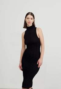 Rochie tricotată fără mâneci, de culoare neagră, cu gât înalt, având un model texturat și o siluetă ajustată, care se termină la jumătatea gambei.