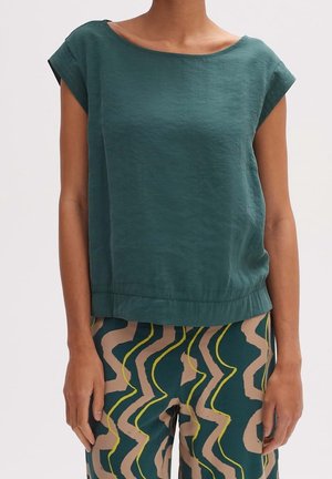 Woman wearing short-sleeve teal top and high-waisted pants with beige, green, and yellow abstract wavy patterns, standing with arms relaxed.