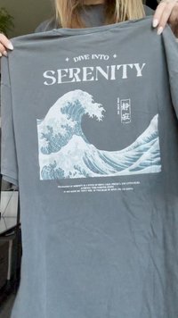 T-shirt de algodão cinza com uma ilustração de onda em azul e branco. O texto diz "MERGULHE NA SERENIDADE" acima e uma descrição abaixo da imagem.