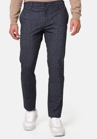 Dunkelblaue maßgeschneiderte Hose aus strukturiertem Stoff mit Knopfverschluss und Seitentaschen, kombiniert mit hellgrauen Sportschuhen.