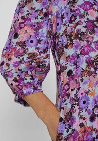 Blommig blus med en ljusblå bakgrund prydd av lila, rosa och orange blommor. Långa ärmar med elastiska manschetter och en mjuk tygtextur.