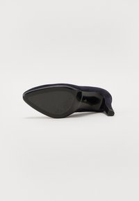 Marineblå semskede høyhelte sko med spiss tå og teksturert såle. Hælen er stiletto-stil, som taper seg til en skarp spiss.
