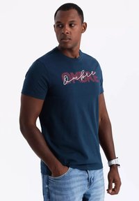 Navy blue cotton t-shirt featuring a "OMBRE" graphic in red and white cursive, short sleeves, and a round neckline, worn with light jeans.