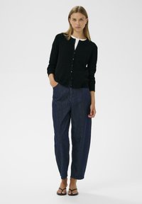 Svart ribbad kofta med rund halsringning och knäppning framtill, kombinerad med lösa blå denimbyxor. Modellen bär svarta sandaler.