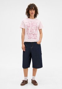Pink cotton t-shirt with humorous graphic illustrations in red. Paired with dark blue, knee-length denim shorts. Brown shoes and white socks.