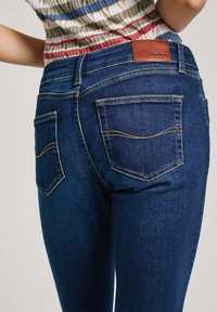 Bakvy av en person som bär mörkblå jeans med två bakfickor och en brun lädervarumärkesetikett, ihop med en ribbad randig topp.