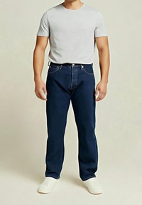 Un homme est debout, portant un t-shirt uni gris clair à manches courtes, un jean bleu foncé et des baskets blanches sur un fond neutre.