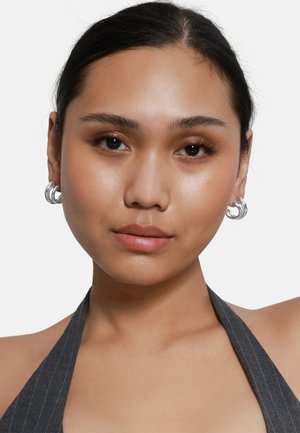 Boucles d'oreilles en argent avec un design tordu et superposé. Le modèle a les cheveux raides et foncés et porte un haut sans manches à rayures foncées.