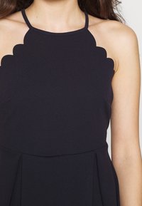 Musta hihaton mekko, jossa on aaltoileva kaula-aukko, valmistettu sileästä kankaasta. Mekossa on laskostettu muotoilu vyötärön ympärillä ja se korostaa siistejä linjoja.