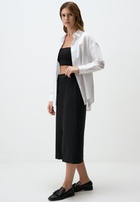 Chemise blanche à boutons sur un crop top noir et des culottes noires, associée à des mocassins noirs avec un détail en nœud. Textures lisses.