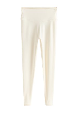 Valgesistist sobiva spordiriietuses ja tossudes naine kohendab oma ülaosa, seistes siseruumis betoonist trepi ja beeži-valge seina lähedal.