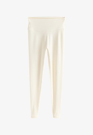 Valgesistist sobiva spordiriietuses ja tossudes naine kohendab oma ülaosa, seistes siseruumis betoonist trepi ja beeži-valge seina lähedal.