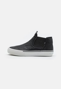Zapatos negros tipo slip-on con material superior texturizado, paneles laterales elásticos y una suela de goma blanca con un sutil acento de franja negra.