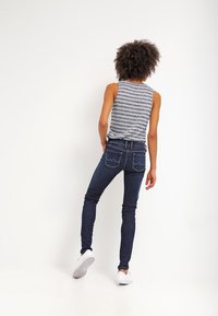 Top sin mangas a rayas grises, jeans ajustados oscuros con sutileza en la costura de los bolsillos traseros, y zapatillas blancas, visto desde la parte de atrás.