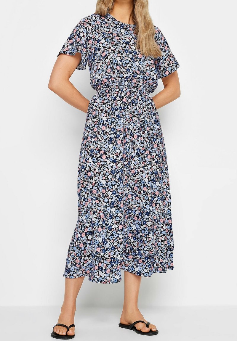 Robe maxi florale en marine avec des motifs de fleurs roses et bleues. Manches courtes, taille élastique, ourlet évasé, portée avec des tongs noires.