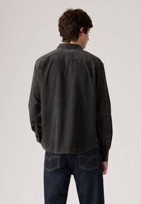 Black denim shirt with a collar, buttoned cuffs, and a curved hem, paired with dark blue jeans featuring contrasting stitching and pocket details.