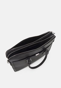 Sorte lædertaske med tre lynlåslommer, glat tekstur, dobbelte håndtag og et logodetalje på den ene side.