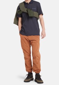 Camisa de polo azul marino, pantalones naranjas de tiro alto, suéter verde sobre los hombros y botas de senderismo negras con suelas de goma, de pie sobre un fondo blanco.