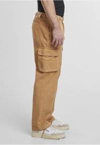 Pantaloni cargo din catifea côtelée de culoare camel, cu croială lejeră, mai multe buzunare laterale și un material texturat. Purtați cu adidași albi.