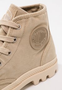 Chaussure montante en toile beige de la marque Palladium avec un patch rond en caoutchouc, des œillets en métal, des lacets et une semelle en caoutchouc texturée.