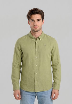 Hombre con cabello castaño que lleva una camisa verde claro abotonada y vaqueros azul claro, de pie frente a un fondo gris liso.