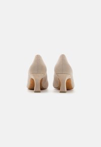 Pumps en suède beige avec un bout pointu et un talon court et large. Le matériau a une texture lisse avec des détails de couture subtils.