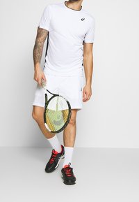 Weißes Sport-T-Shirt mit schwarzen Akzenten, kombiniert mit weißen Shorts. Hält einen Tennisschläger, trägt schwarze Schuhe mit roten Schnürsenkeln und weiße Socken.