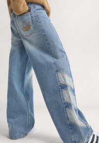 Ljusblå denimjeans med vida ben, prydda med en broderad logga på bakfickan och diagonala paneldetaljer längs sidorna.