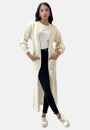 Lungo cardigan crema con polsini a coste e tasche, indossato sopra una maglietta bianca e pantaloni neri, con una vestibilità rilassata e una texture morbida.