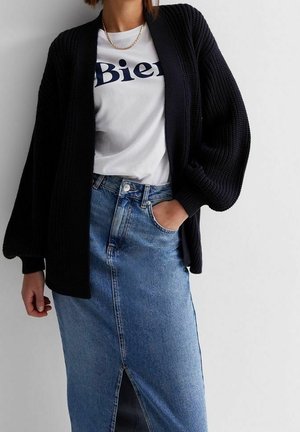Personne portant un t-shirt blanc "Bien", un cardigan en maille épaisse noir, un collier chaîne doré et une jupe en denim taille haute avec une fente à l'avant.