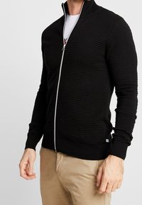 Černý svetr na zip s vroubkovanou texturou, vysokým límcem a kontrastním bílým zipem. Přiléhavý střih, nošený přes bílé tričko.