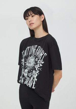 T-shirt nera oversize con un design grafico argento che presenta un sole, elementi floreali e la frase "La fortuna aiuta gli audaci."