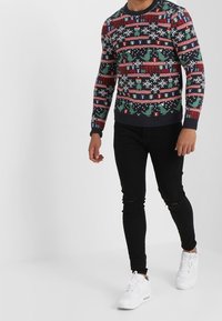 Pull de Noël tricoté avec un motif de flocon de neige et de dinosaure vert et blanc, agrémenté de rayures rouges et bleues. Associé à un jean skinny noir et des baskets blanches.