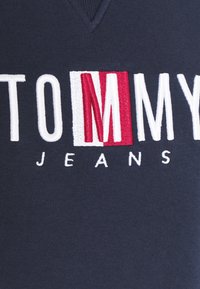 Tissu bleu foncé avec des lettres blanches brodées formant "TOMMY" et des lettres blanches plus petites en dessous formant "JEANS,", avec "M" mis en évidence en rouge et blanc.