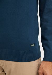 Tumesinine kudum-sviiter ribatud servade ja mansettidega; silt alumises vasakus nurgas, sile tekstuur, sobiv disain. Ühendatud beežide pükstega.