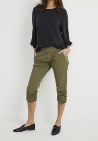 Svart långärmad skjorta, olivgröna cropped byxor med rynkade ärmslut och svarta loafers; avslappnad passform, med fickor och synlig sömnad.