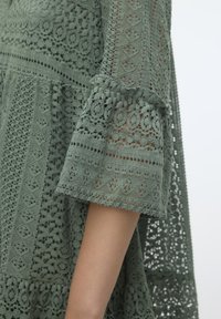 Robe verte en dentelle ornée de motifs complexes, avec des manches évasées et un design superposé à mailles ouvertes, mettant en valeur un tissu texturé.