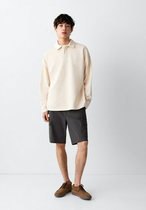 Jonge man staand, gekleed in een crèmekleurig poloshirt met lange mouwen, donkergrijze cargoshorts en bruine veterschoenen, tegen een effen muur.