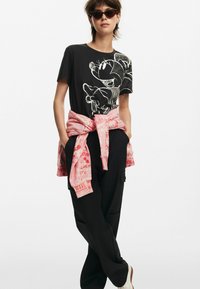 Camiseta negra con gráfico de dibujos animados blanco, combinada con pantalones cargo negros y un sudadera rosa atada a la cintura. Las gafas de sol completan el conjunto.